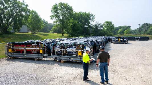 Two men stand in a lot at Argonne National Laboratory next to several rows of large, complex magnets covered in black tarps, stretching back to the edge of the yard