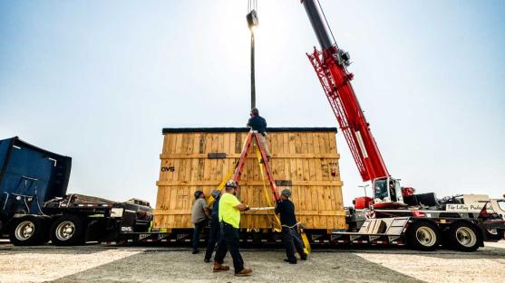 Two workers look on as a large red crane moves an enormous wooden crate from the bed of a truck. 
